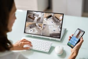 A woman using a smartphone app to view live footage from a smart home security camera on her computer screen. This image illustrates the benefits of smart homes, specifically remote monitoring capabilities.