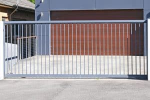 Automated entrance gates in grey steel, lead to a modern house with a matching grey exterior and brown garage door.