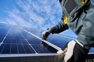 Technician performing solar panel maintenance on a residential solar PV system, wearing protective gear and working under a clear blue sky.