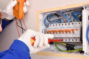 Electrician in protective gear using a multimeter to inspect an electrical circuit breaker panel, demonstrating steps to troubleshoot and prevent flickering lights in a home.