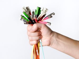 Hand gripping colorful fiber optic cables with various connectors against a white background, illustrating how to configure fiber optic network.